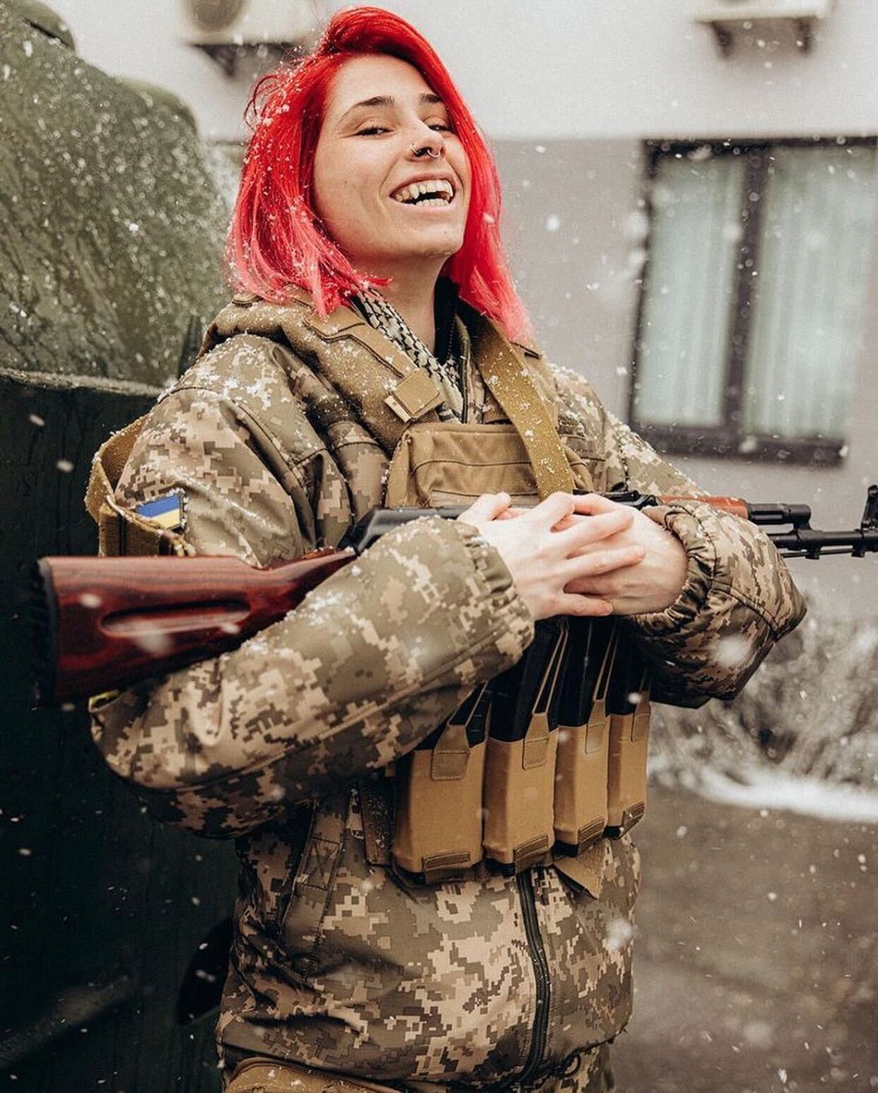 photo of Ukrainian soldier with dyed pink hair and a gun, smiling as snow falls