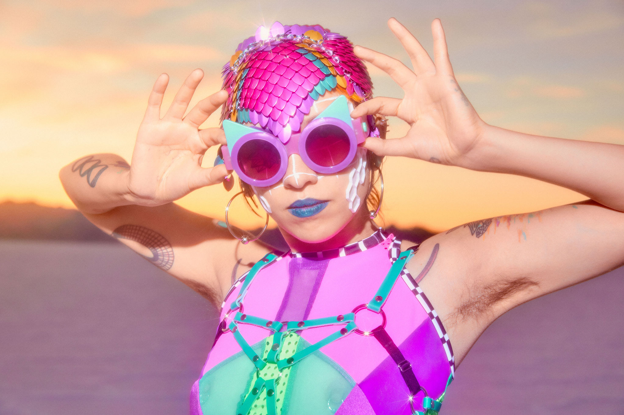Photo of Sky Cubacub, a Filipinx nonbinary person modeling a skin tight cap made of bright metallic pink, teal, and gold fish scales, teal lipstick, round pink fashion sunglasses, hoop earrings, and a spandex pink and teal halter top with a matching harness.