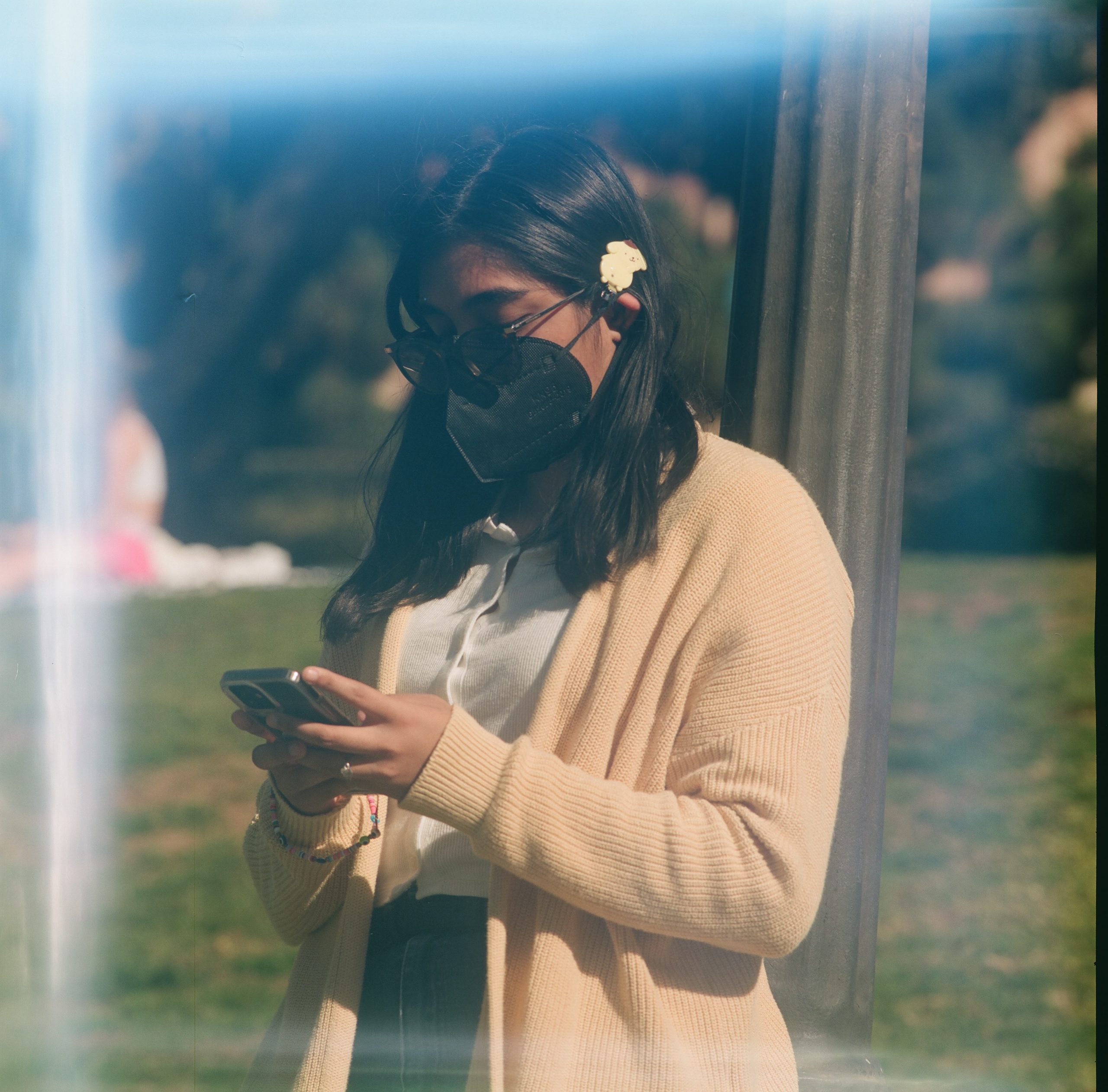 photo of a Brown nonbinary person with shoulder-length black hair, glasses, a yellow sweater over a white button down, a black face mask, and black pants. they stand in the UCLA botanical gardens on their phone with a flower tucked behind their ear.