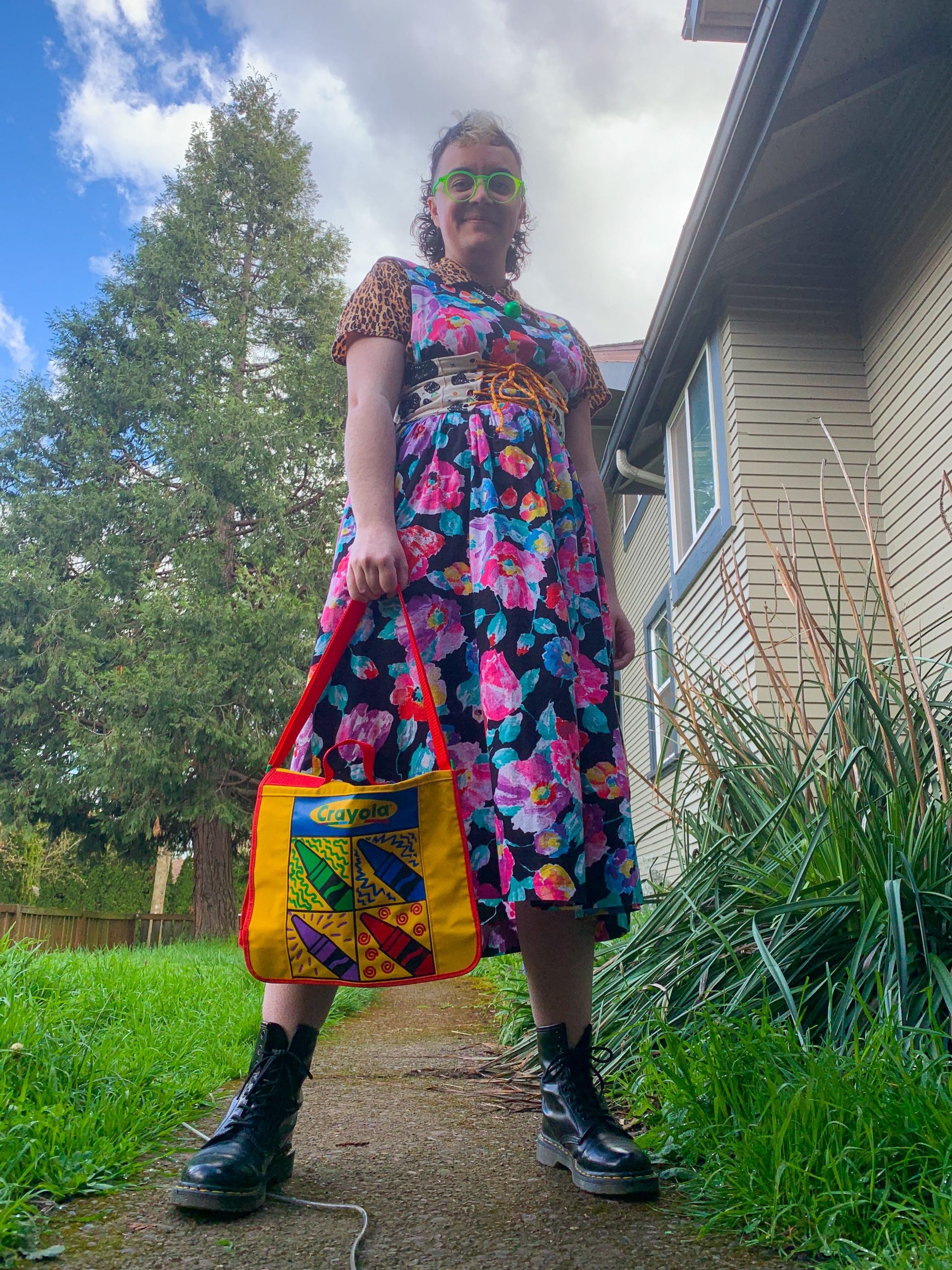 Photo of Juniper, a white autigender person with a brown and blond mullet, neon green glasses, a colorful short sleeved dressed patterned with flowers, a colorful crayola themed tote bag, and black Dr Martens standing powerfully in its backyard.
