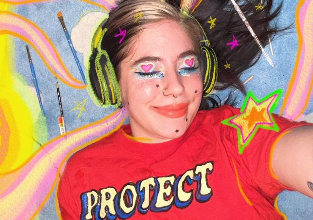 photo of Rhiannon, a white nonbinary person with colorful makeup and a red Protect Trans Youth shirt