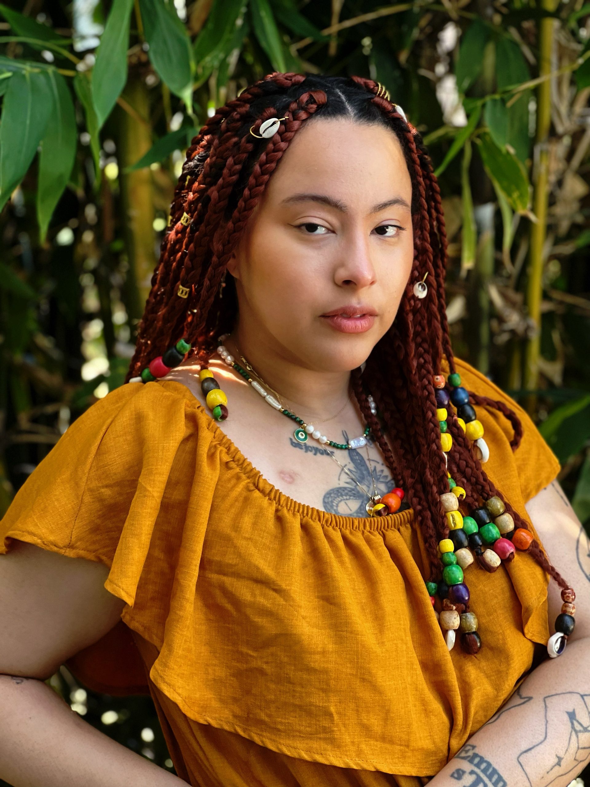 photo of Walela Nehanda, a Black nonbinary person with dark braids threaded with colorful beads and shells, a beaded necklace, a deep yellow layered top, and tattoos. they stand in front of green foliage with a powerful expression