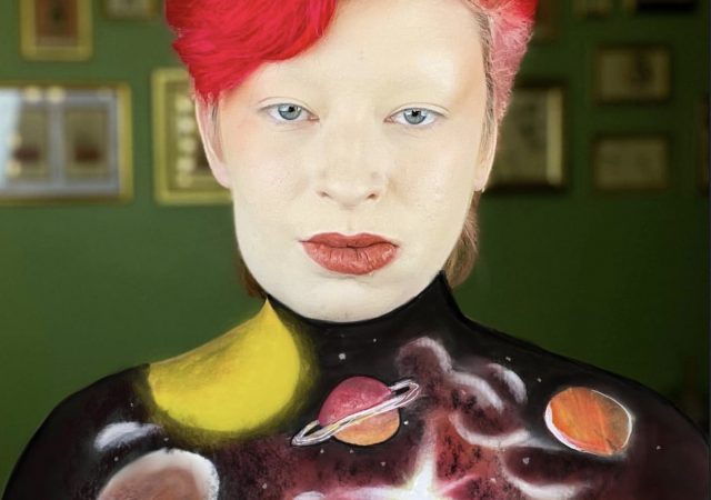 photo of The Mother Birdie, a white nonbinary person with short, bright red hair, bold red lips, and their chest painted with realistic planets and stars