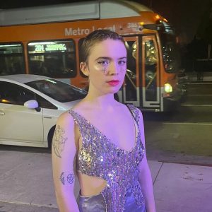 Photo of Layla, a white genderfluid person with buzzed dark hair, a sparkly silver dress, and matching glittery makeup.