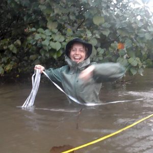 Photo of Zoë, a white woman with wet blonde hair tucked into a bulky raincoat. She is laughing while wading through a river next to some foliage, holding a line of rope. 