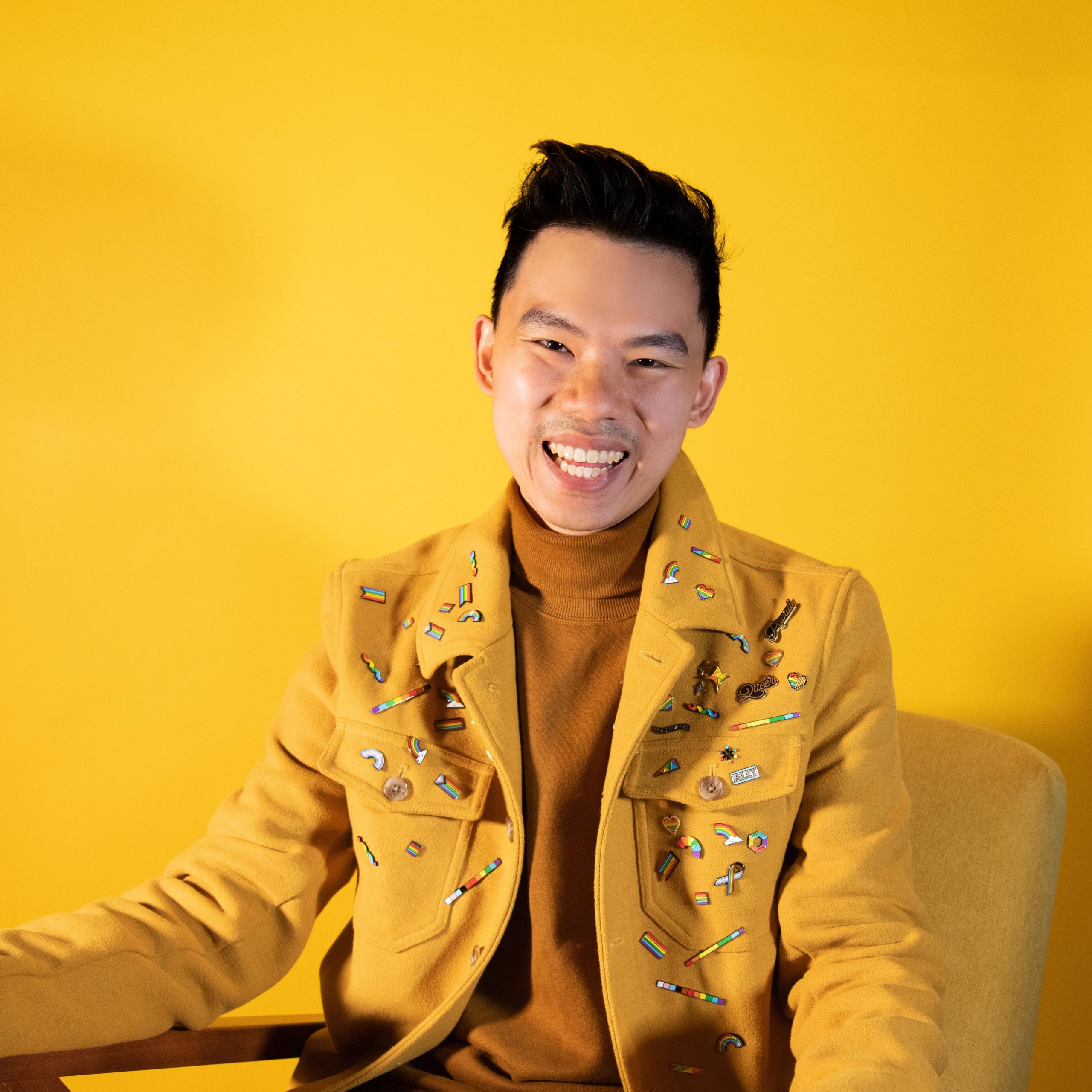 photo of Zar, a smiling Singaporean-British man with short black hair. He is wearing a yellow blazer with tons of pride pins over a darker yellow sweater. behind him is a yellow background.