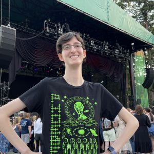 Photo of Christopher, a white genderqueer man with short brown hair, glasses, and a black shirt with a neon green alien graphic. He smiles with his hands on his hips in front of a stage.