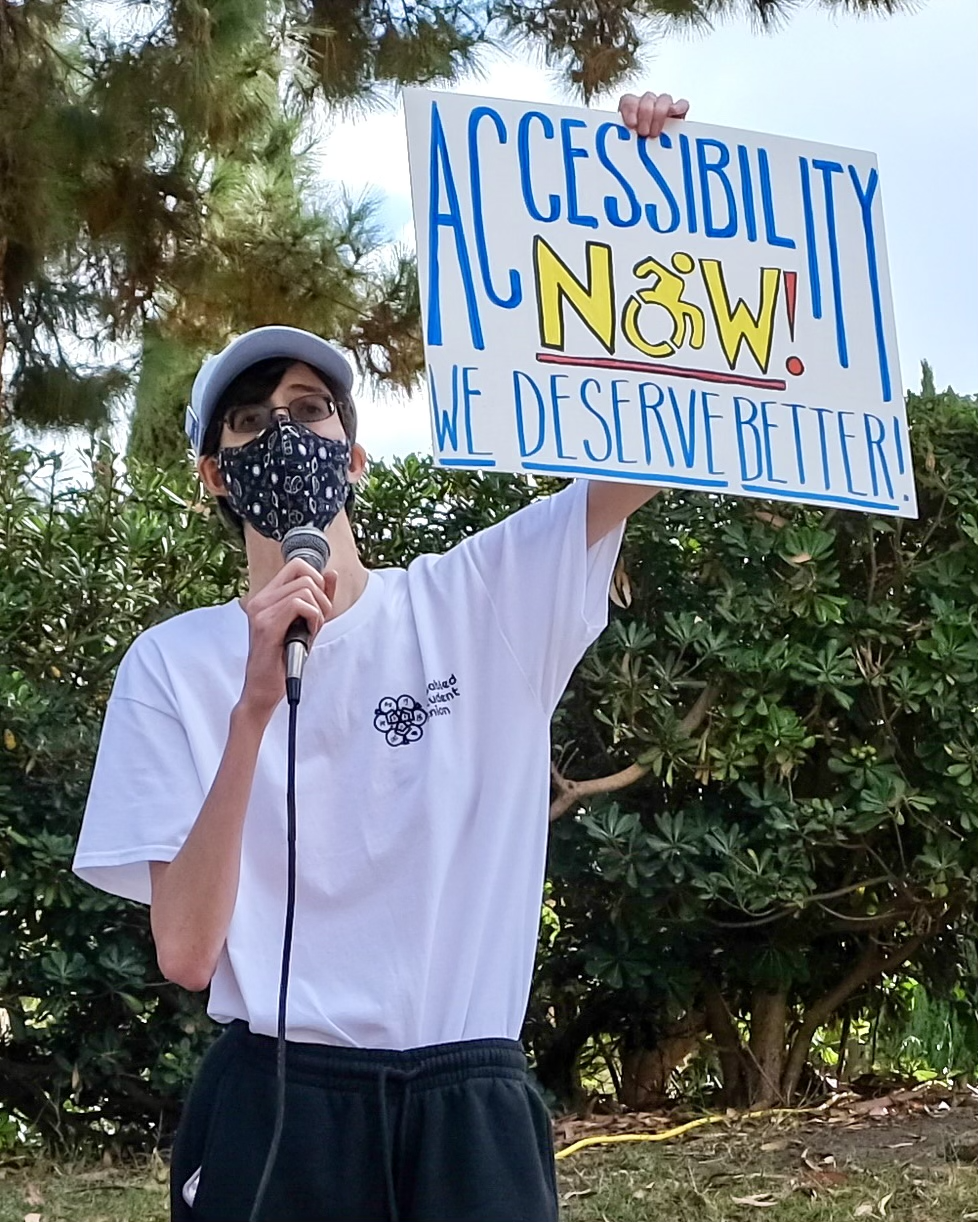 photo of Christopher, a white genderqueer man with short brown hair tucked under a light blue baseball cap, glasses, a black mask, a white Disabled Student Union shirt, and black pants speaking into a microphone and holding up a protest sign that reads "Accessibility Now! We Deserve Better."