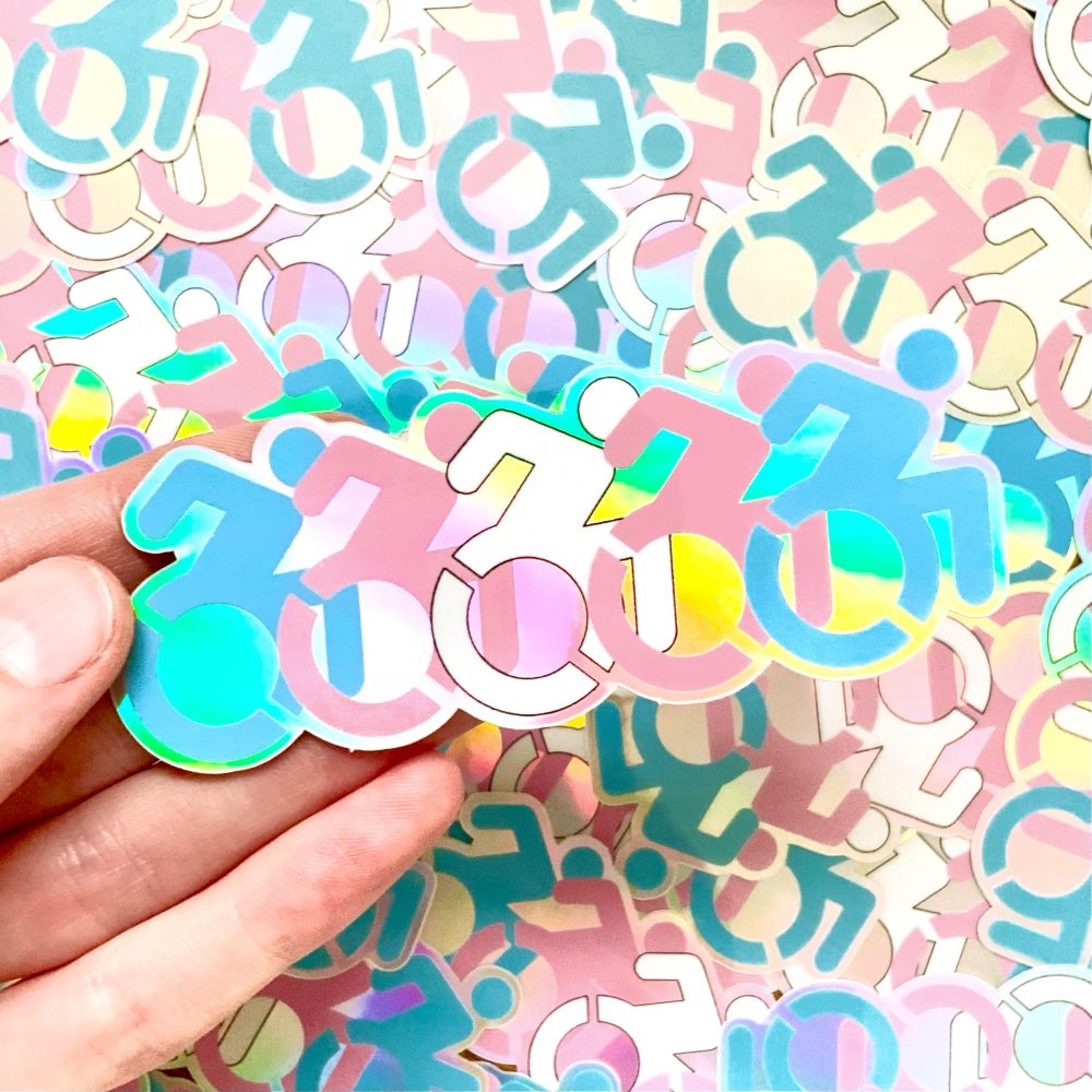 photo of a pale hand holding a holographic sticker in front of a pile of the same design. the design features 5 disability wheelchair symbols with each colored to match a stripe of the transgender pride flag.