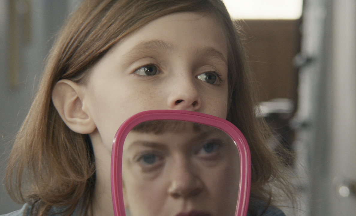 still from the film "Homebody" showing the young blonde boy Johnny looking over a mirror at Melanie, his babysitter, reflected in the glass