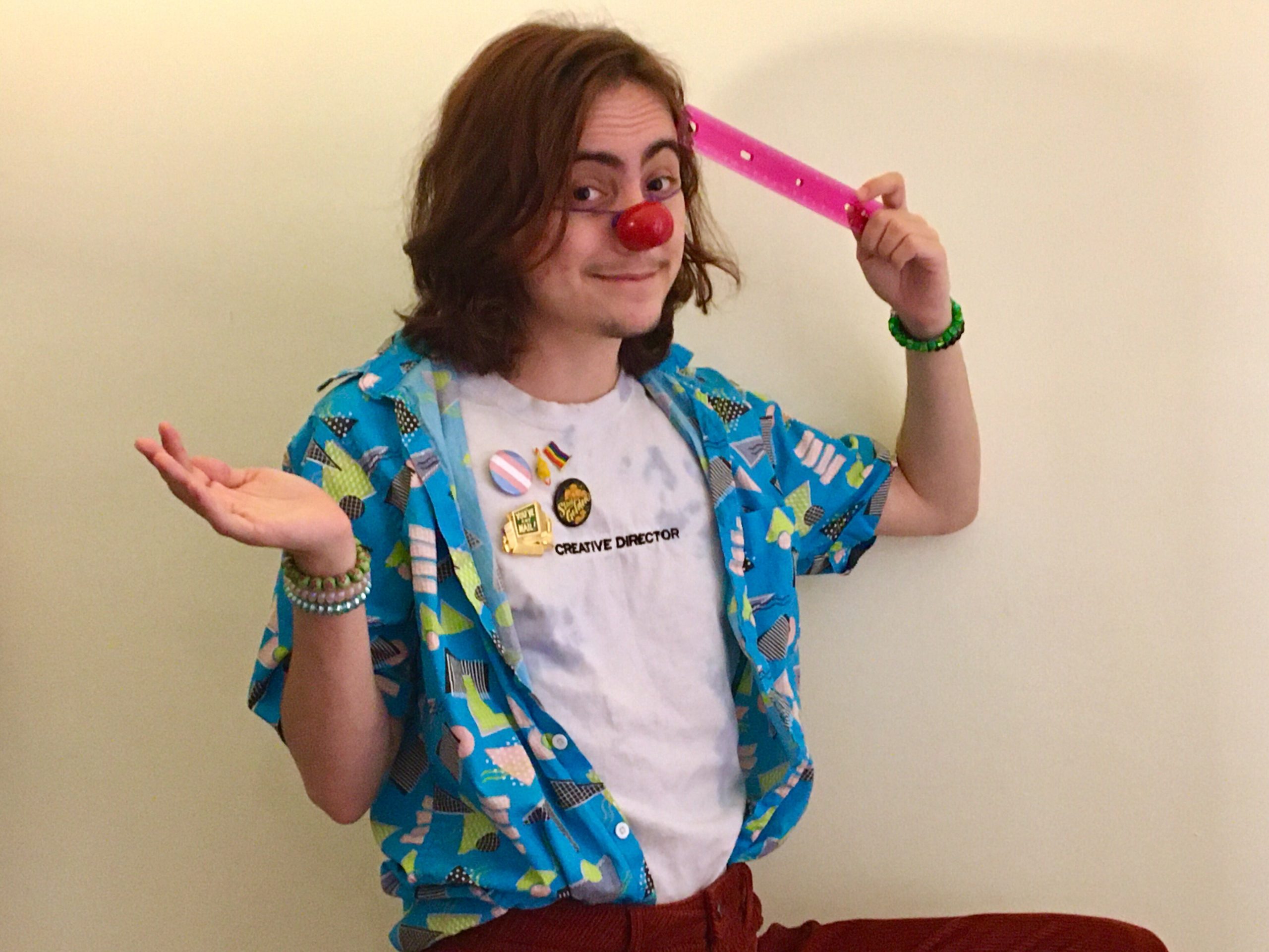 photo of Emmet Abrams, a white transmasculine person with shoulder length light brown hair, an open patterned shirt over a white top with pride pins, and a red clown nose.