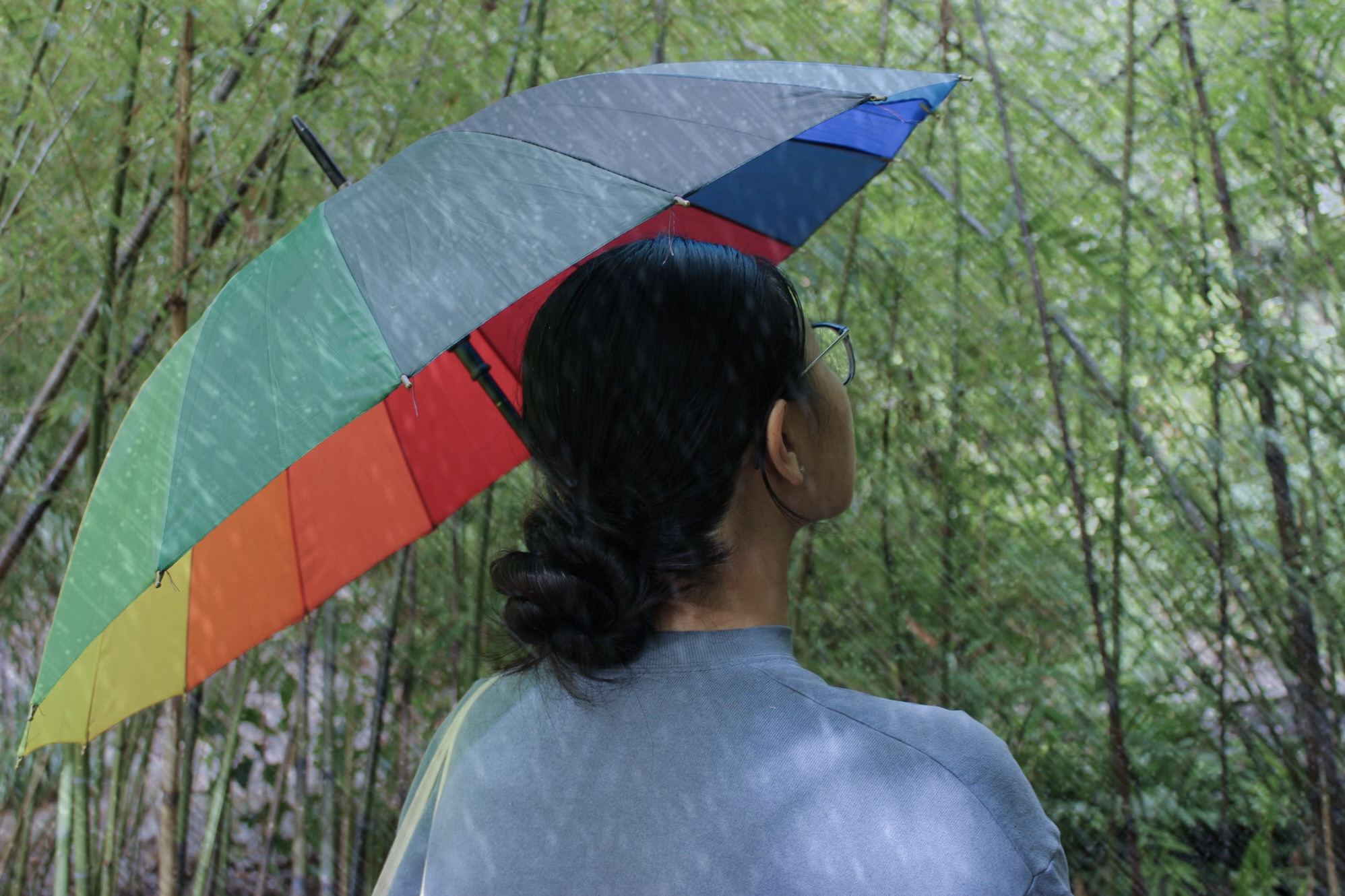 photo of a black haired woman standing under a rainbow umbrella as it rains