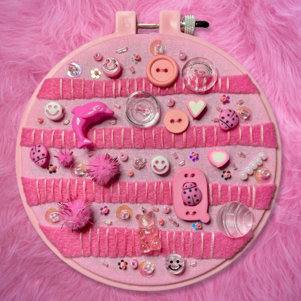 photo of an embroidery hoop with a design featuring thick and textured pink stripes under dozens of pink, clear, and white charms and buttons of different shapes and sizes.