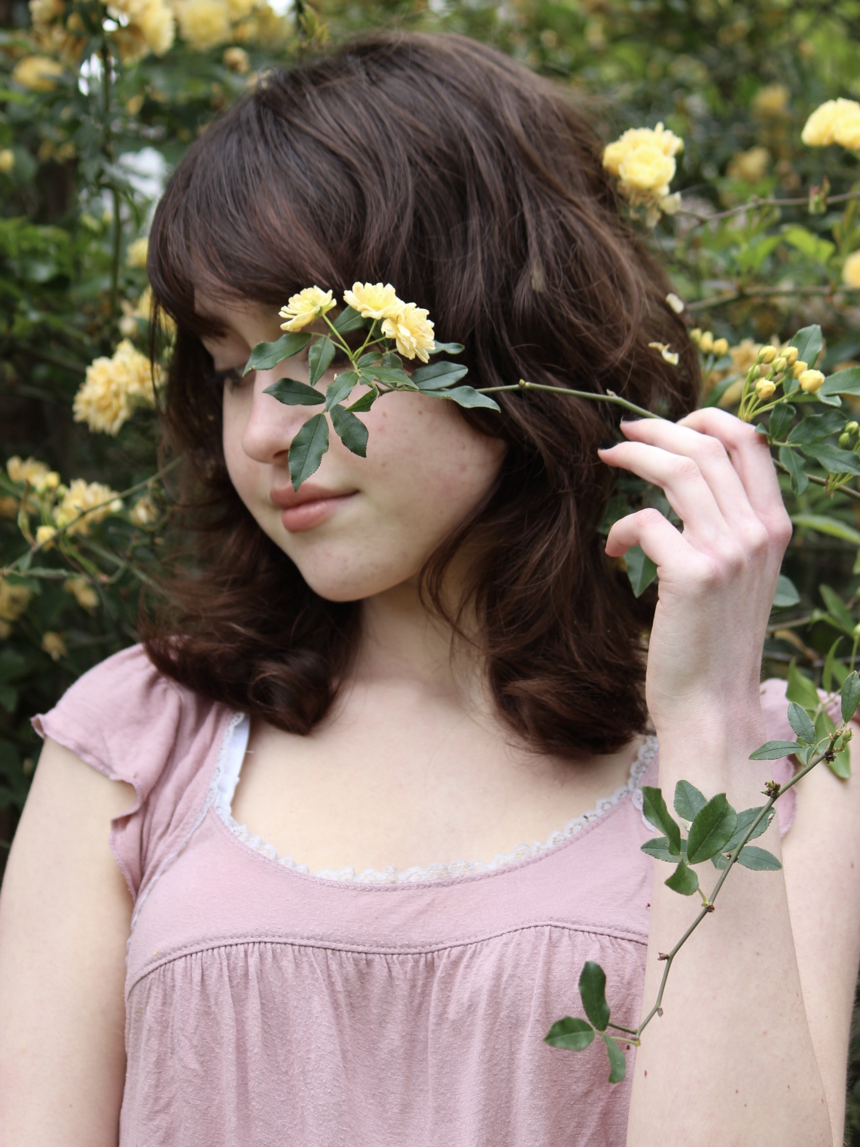 photo of a brown haired girl holding a flower in front of her face, partially hiding a smile