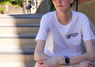 A photo of Christopher, a white genderqueer man, sitting on stairs on UCLA campus. He has a serious expression and wears tinted glasses, a watch, and a white shirt reading, "Disabled Student Union."