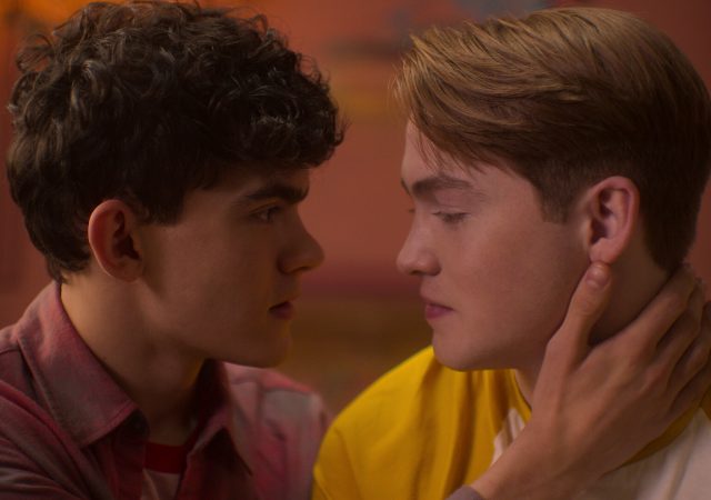 A photograph of Nick and Charlie staring lovingly at each other from Netflix's "Heartstopper". Charlie is a young white man with black hair and a red plaid shirt. Nick is a young white man with blonde hair wearing a yellow shirt. Charlie cups Nick's face.