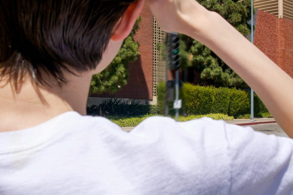 A photograph of Christopher, a white genderqueer man, from behind. He holds his hand up to his head and looks out to the side of a building.