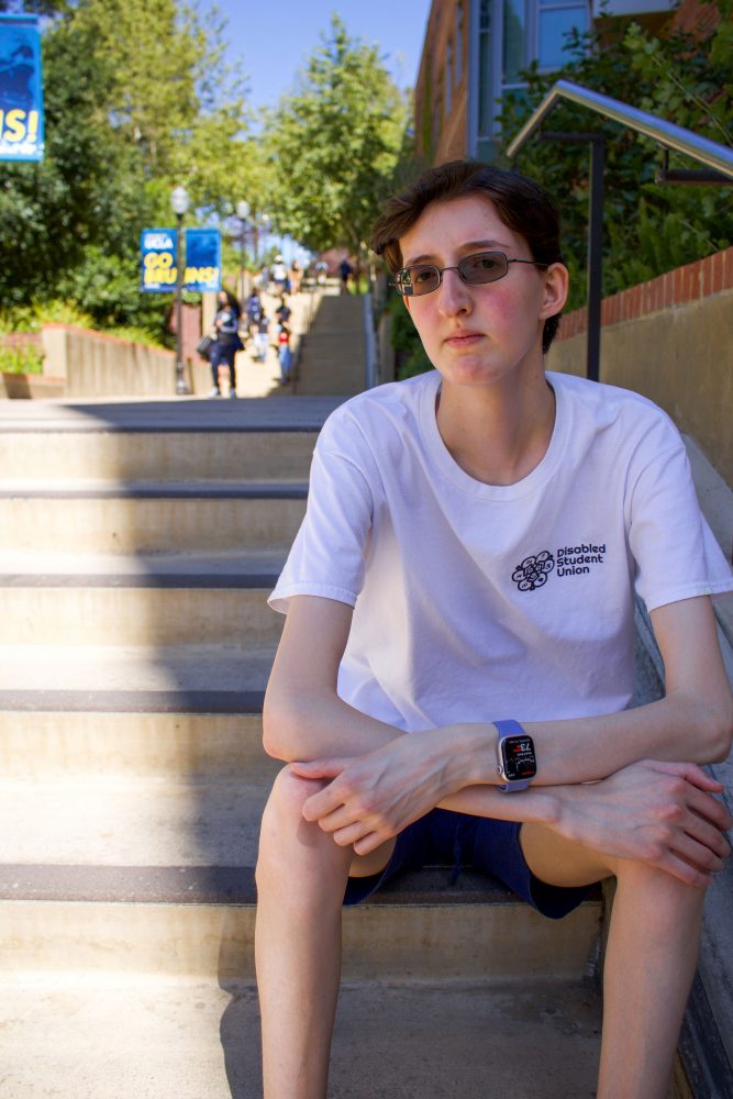 A photo of Christopher, a white genderqueer man, sitting on stairs on UCLA campus. He has a serious expression and wears tinted glasses, a watch, and a white shirt reading, "Disabled Student Union."
