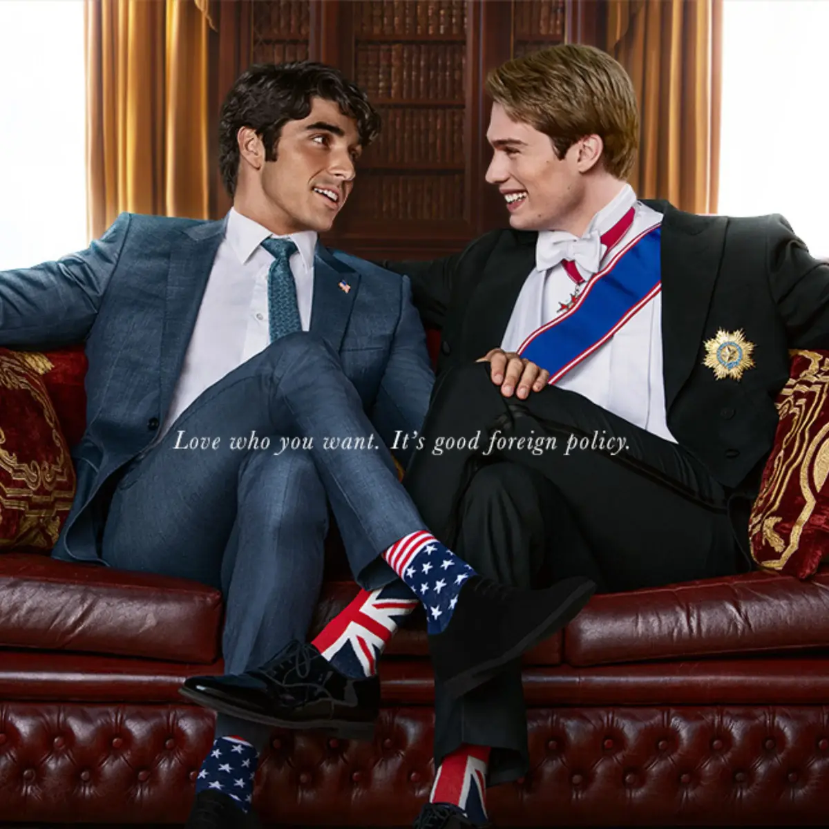 A photograph of a smiling Alex, a Latino man with light brown skin and black hair wearing a gray suit and American flag socks, sitting on a couch with a smiling Henry, a white man with blonde hair, wearing a black suit, blue slash, and Union Jack socks. The photograph reads, "Love who you want. It's good foreign policy."