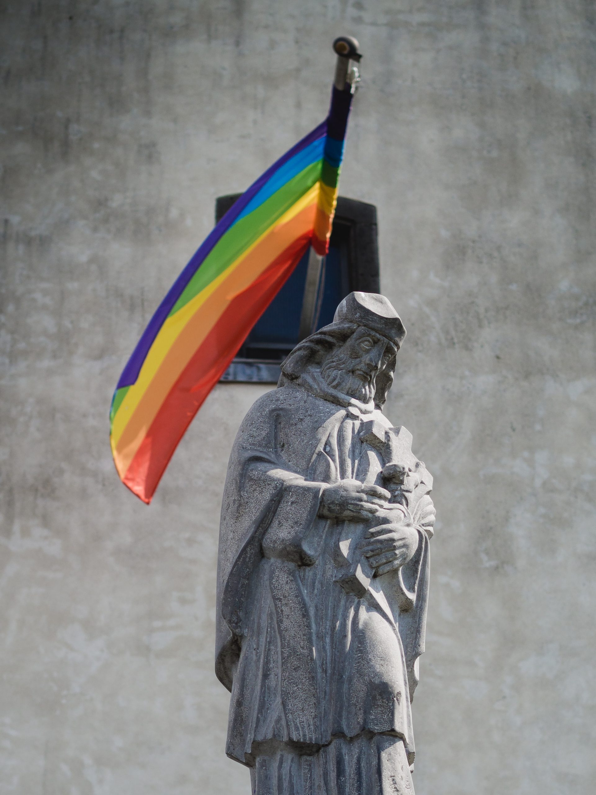A photograph of a stone Catholic saint statue. The saint is a bearded man wearing a hat and robes and carrying a cross. Behind him, there is a stone wall with a window. A rainbow flag juts out of the window.