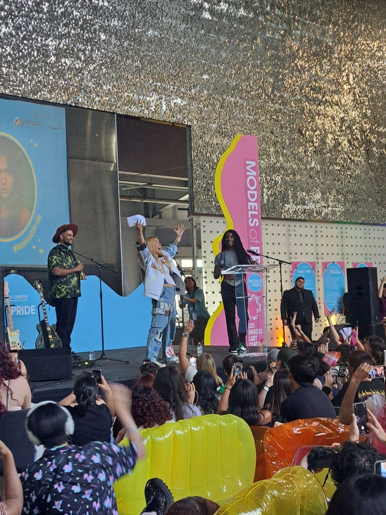 also for monday article: A photograph of Hayley Kiyoko, a white and Japanese woman with tan skin, in a denim outfit on a stage. She has her arms raised in the air. On stage with her are a Black masc person in a brown hat and green shirt and a Black person with long black hair in a gray outfit. Behind her, the wall has a blue wave design. At the bottom of the stage, the audience members raise their hands excitedly.