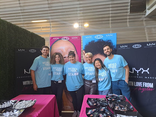 A photograph of the NYX Cosmetics booth. It has a black background and two pink tables. The six staff members wear blue Models of Pride staff shirts.