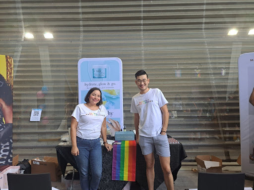 A photograph of the Philosphy booth, a black table with a rainbow runner in the middle. Two people stand in front of the booth: a femme person with light skin and a short black bob and an Asian masc person with short black hair and glasses. Both of them wear white shirts that say, "Perfect. Marc Jacobs." in a rainbow gradient. Behind their booth is a stand-up banner that has various skincare products on it.