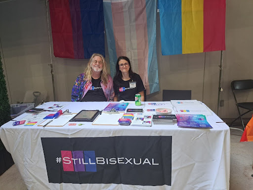 A photograph of the #StillBisexual booth. Seated behind the booth are two white people wearing black #StillBisexual shirts with the bi flag colors behind the word "Still". One is an older masc person with long blonde hair, a white beard, and glasses. The other is an older femme person with long black hair and glasses. Behind them, are the bi flag, the trans flag, and the pansexual flag hanging from the ceiling. Their booth has a white tablecloth and various fliers atop it.