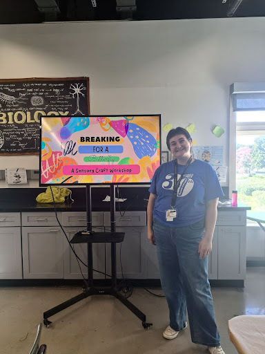 A photograph of MFT trainee Jules Wolfe, a white person with short black hair wearing a blue Models of Pride 30th Anniversary shirt and jeans. They stand beside a TV which says, "Breaking for a Butterfly: A Sensory Craft Workshop" on a colorful PPT slide.