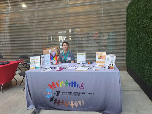A photograph of a masc person with light skin and brown hair wearing a gray shirt, smiling. They are seated at the Burbank Community YMCA booth. The booth has a gray tablecloth and has various papers propped up on top of it.