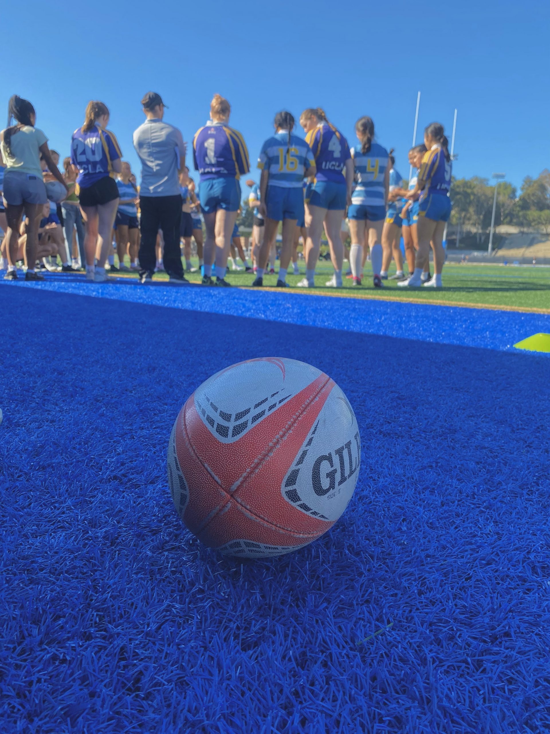 A photograph of a white and orange rugby ball on blue artificial turf. In the background are UCLA rugby players.