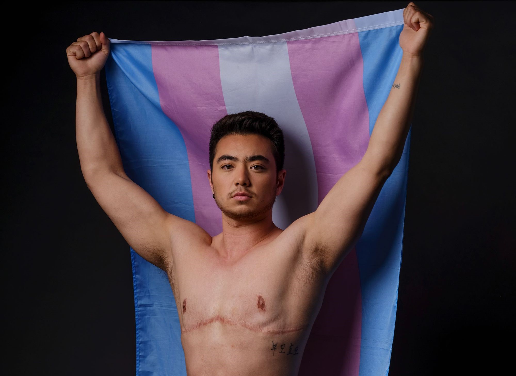 A photograph of Schuyler Bailar, an Asian trans man with light skin, black hair, and black facial hair. He is shirtless with visible top surgery scars, and he holds the trans flag above his head; the flag streams out behind him.