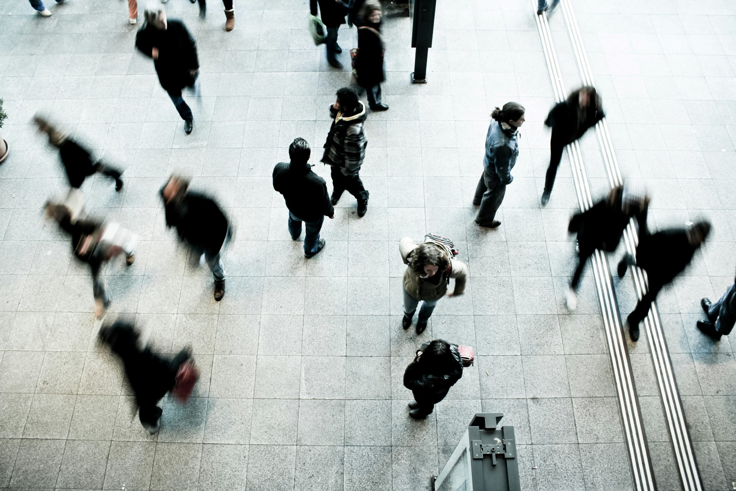A photograph taken from an aerial view of blurry people walking in various directions. The ground is composed of gray stones.