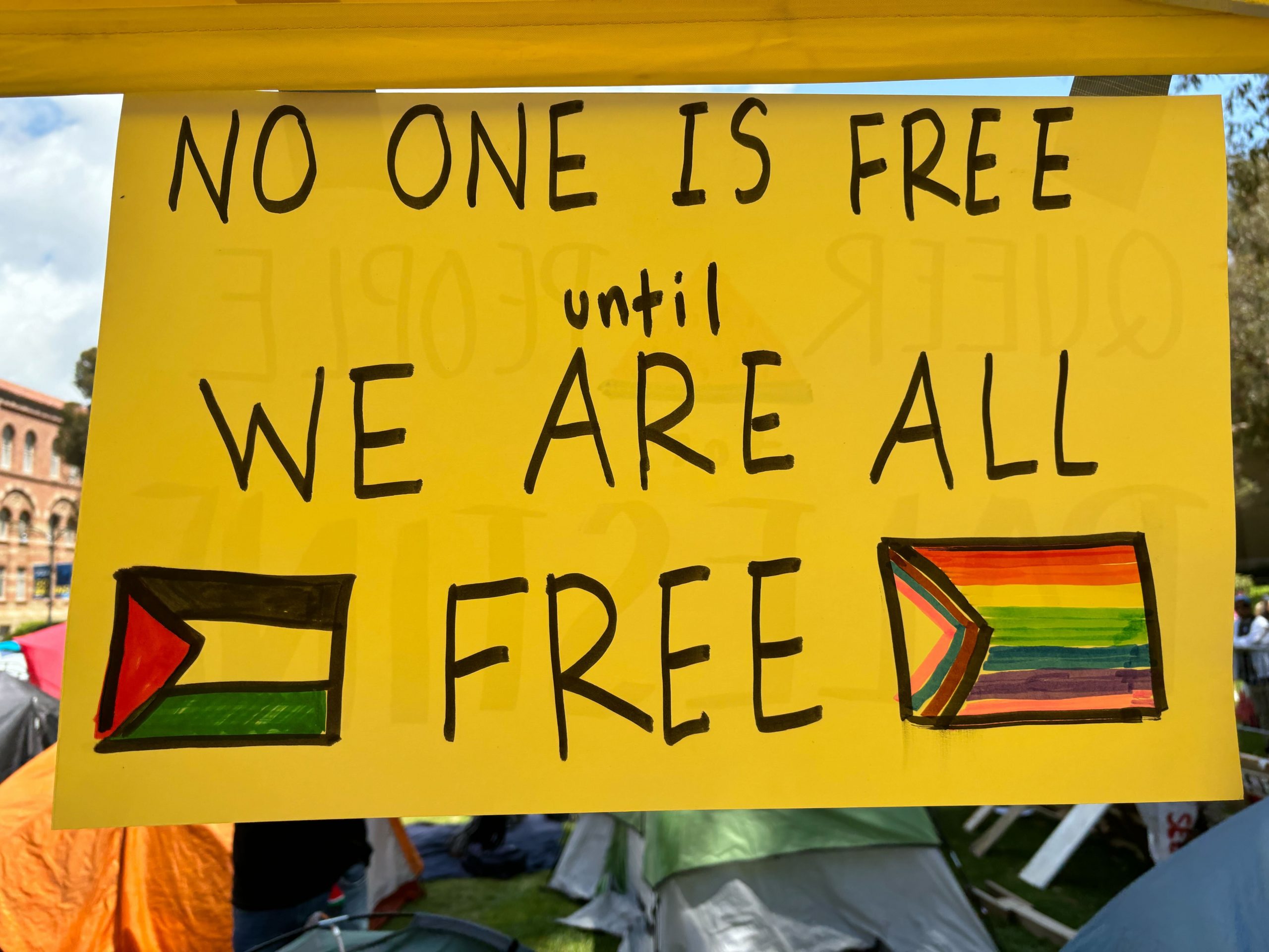 A yellow paper sign hung from a tent at UCLA’s Palestinian Solidarity Encampment. Handwritten black text reads “No one is free until we are all free” between a Palestinian flag on the left and a Progress Pride flag on the right.