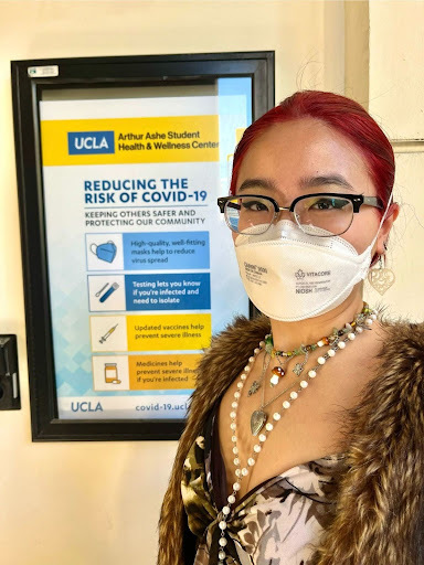 A photograph of Gwen, an Asian woman with red hair, glasses, and light skin. She's wearing a shirt with fur lining and multiple necklaces. Gwen stands in front of an informational poster entitled "Reducing the Risk of COVID-19."