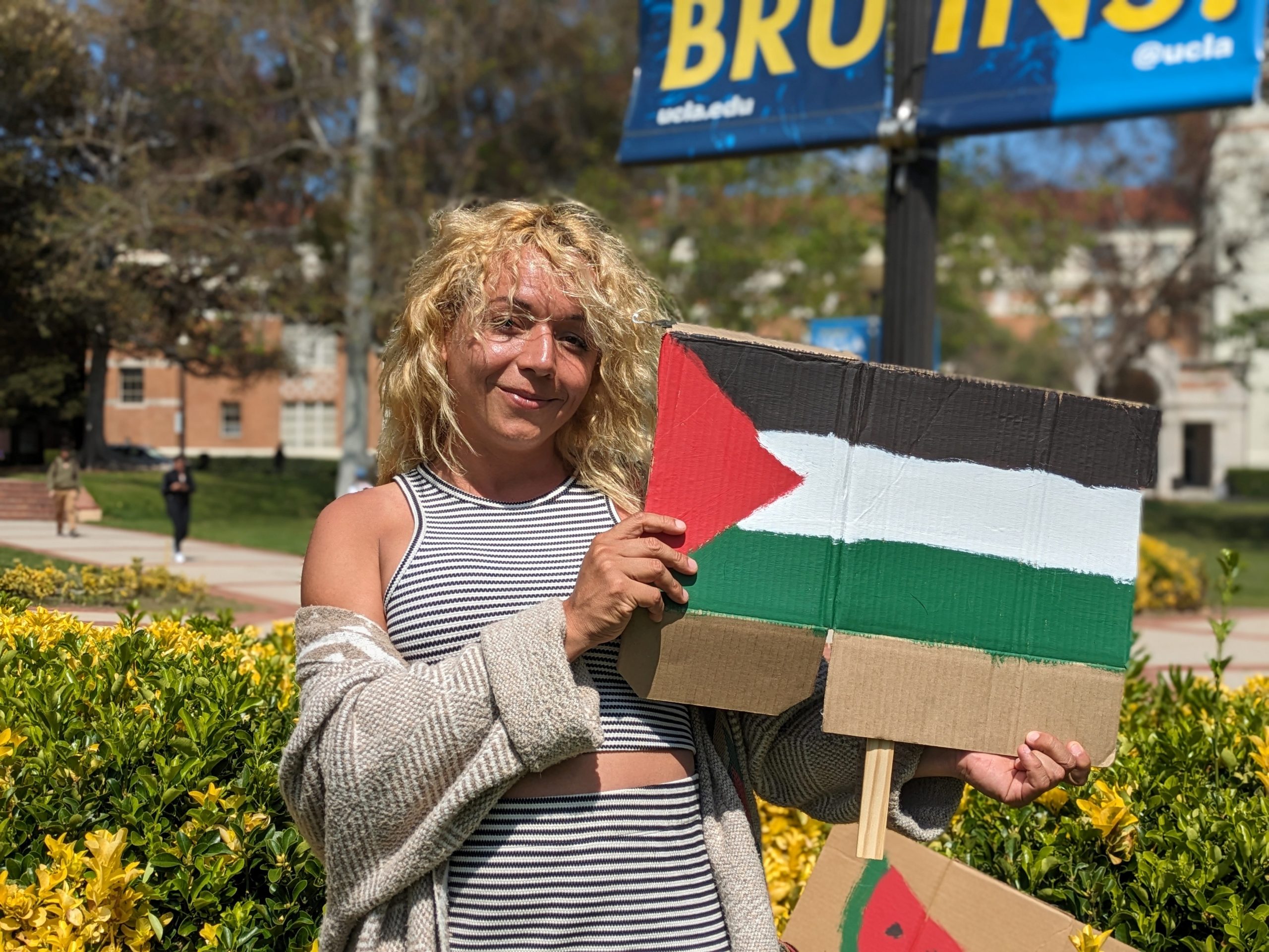 A photograph of Daniela Maldonado Salamanca, a Brown woman with wavy blonde hair, wearing a matching two-piece black-and-white striped tank top and skirt set. She smiles as she holds a cardboard sign with a painted Palestinian flag. Behind her is Dickson Court and a Bruins sign hanging on a lamppost.