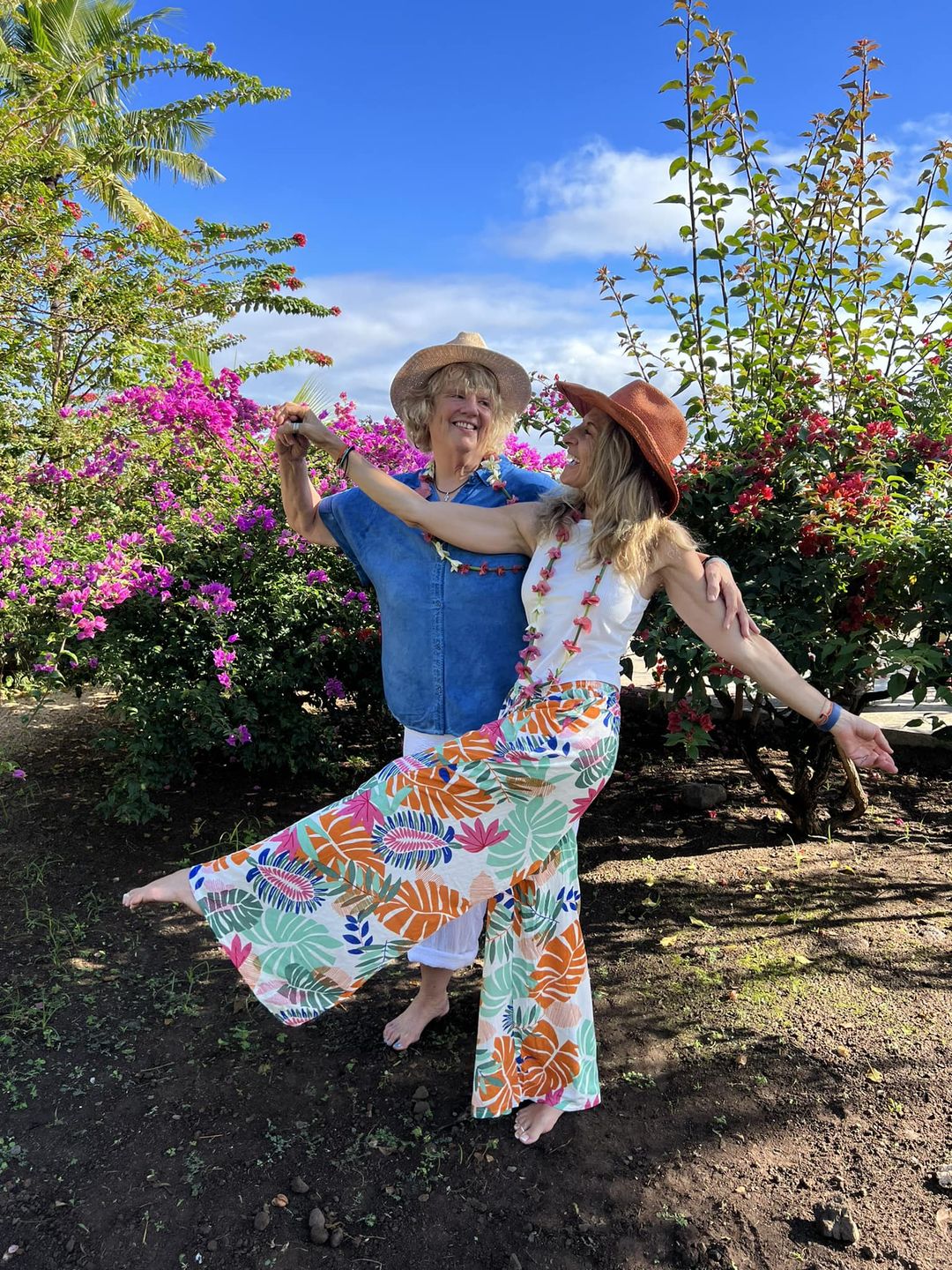 Caryn and Aly, Sarah's aunts, dance barefoot in a garden. They are older white women wearing summery clothing. Both are smiling widely.