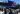 A crowd of anti-RIMPAC protestors with banners and flags gather in front of a U.S. Navy aircraft carrier.