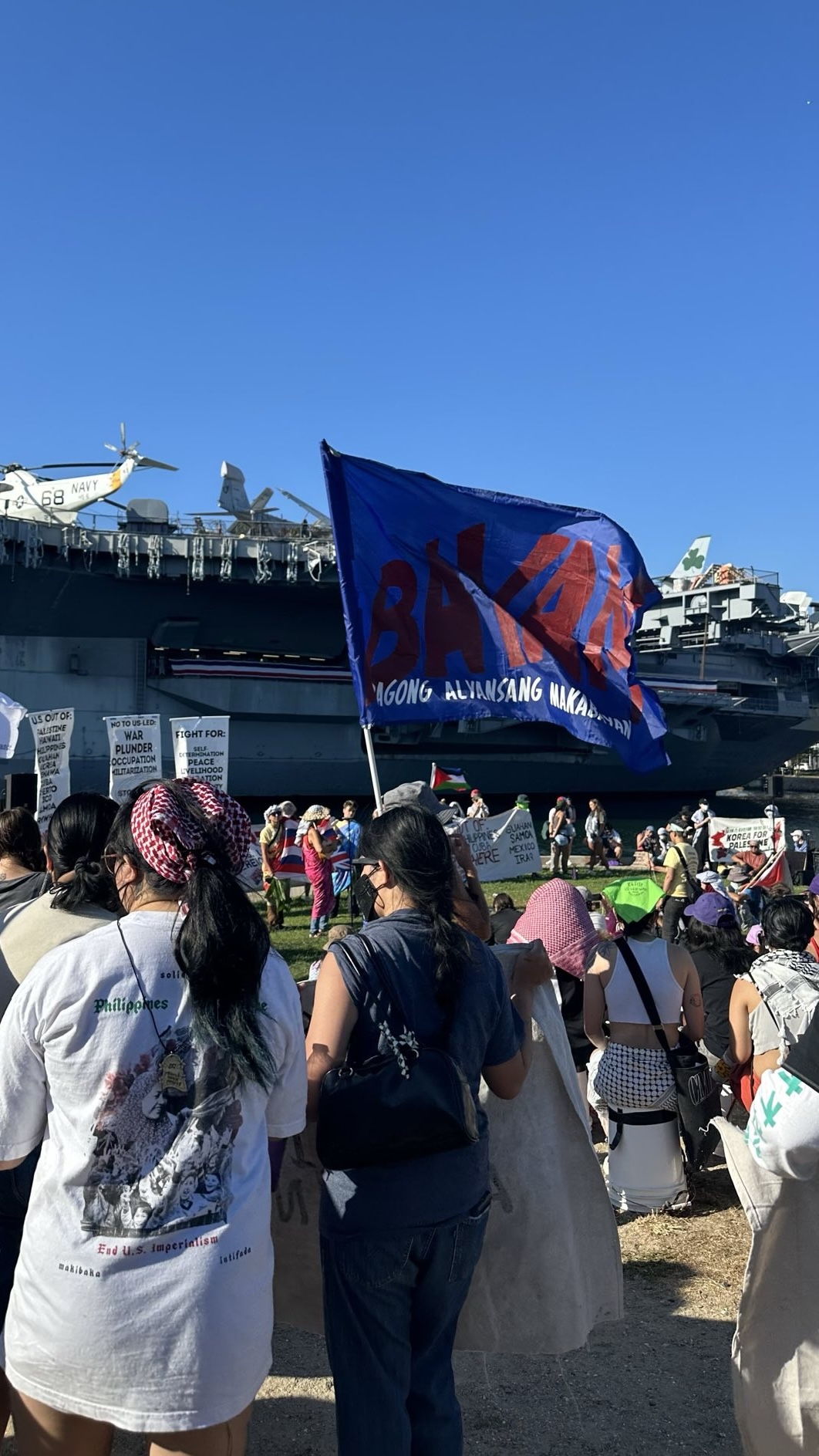 A crowd of anti-RIMPAC protestors with banners and flags gather in front of a U.S. Navy aircraft carrier.