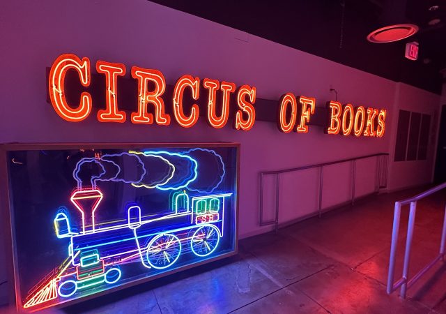 A neon sign reading Circus of Books above a neon sign of a train.