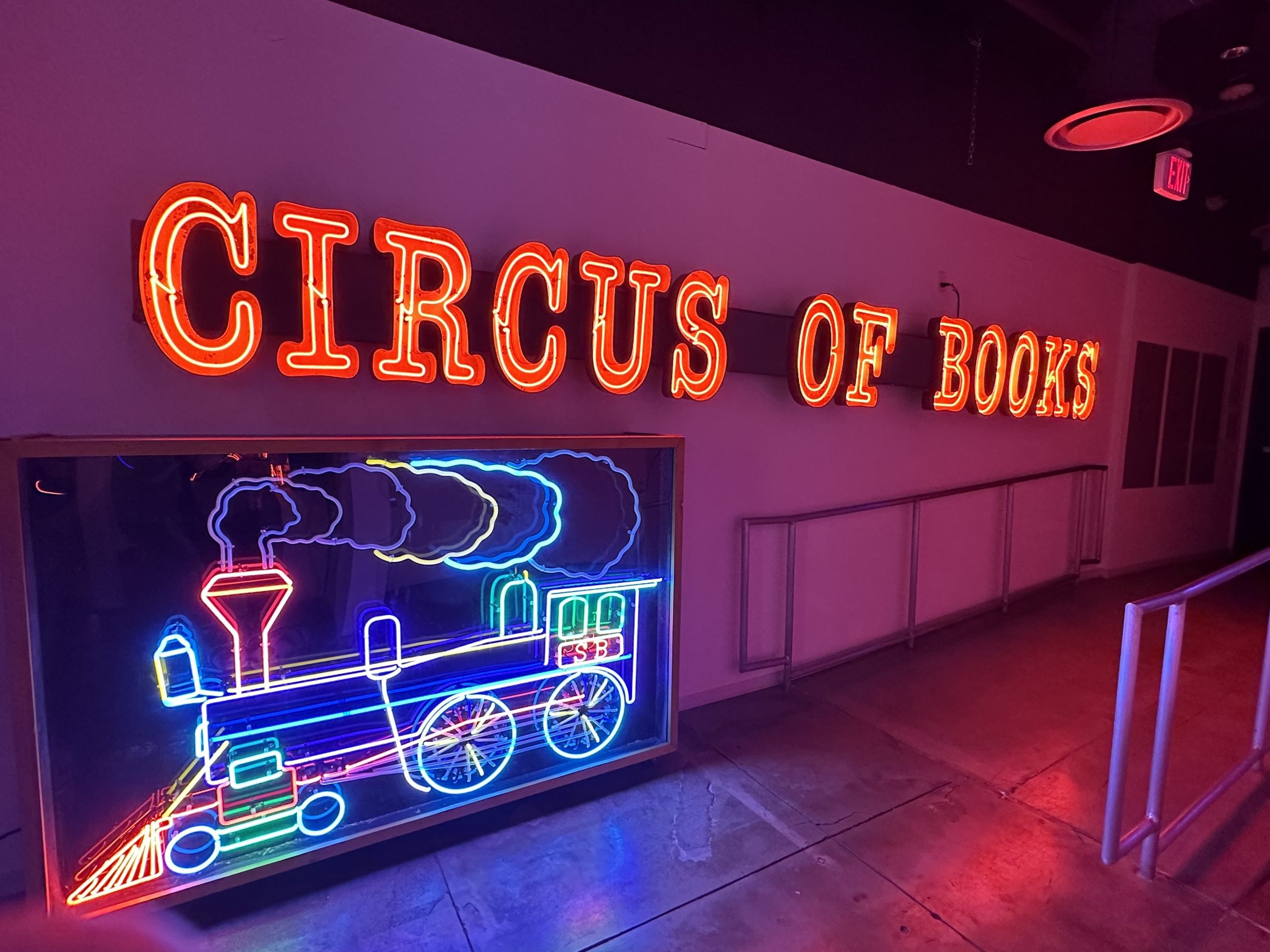 A neon sign reading Circus of Books above a neon sign of a train.