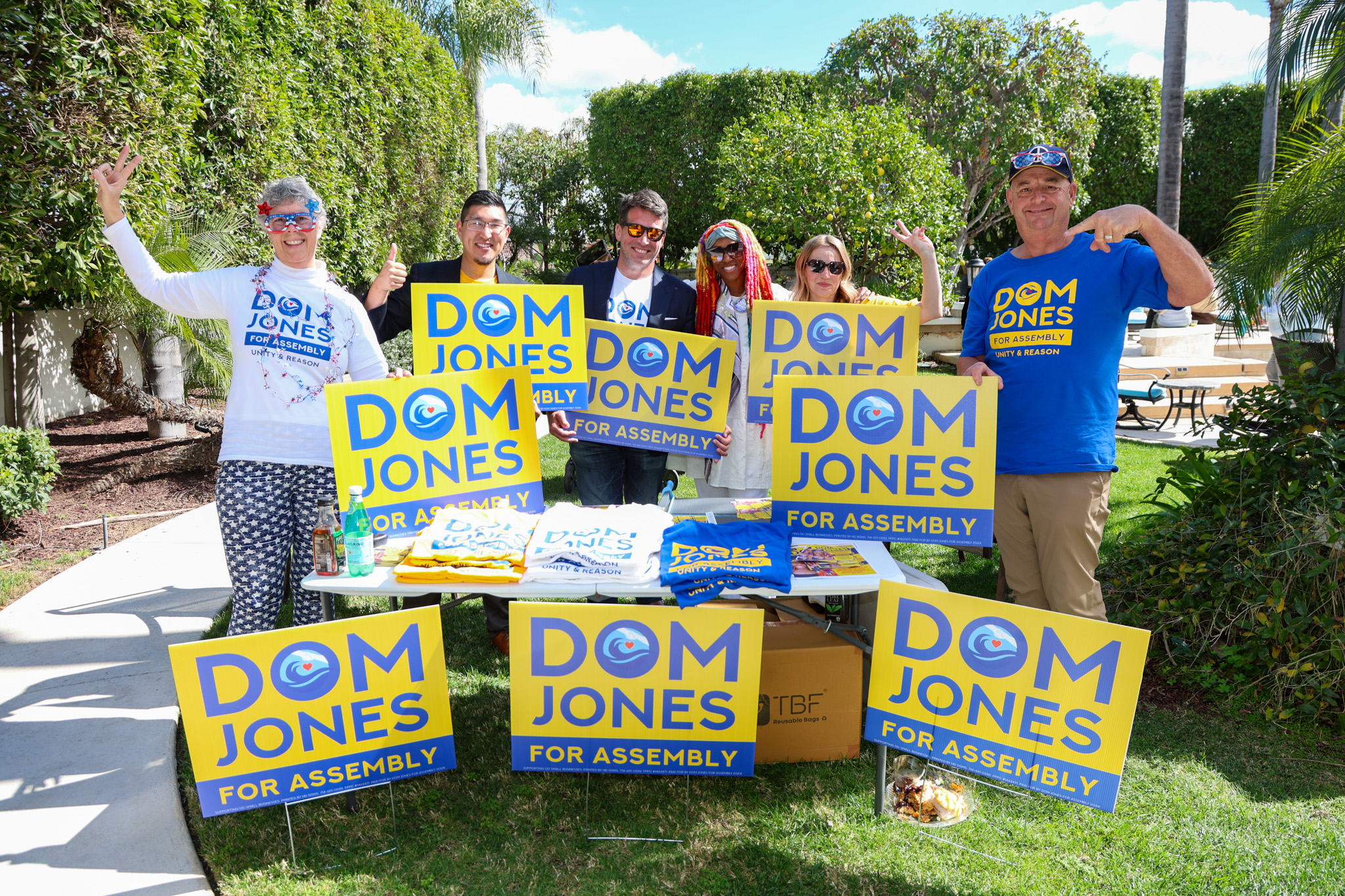 A group of diverse people standing on a lawn holding Dom Jones for Assembly signs. Among them is Dom Jones, a Black woman with colorfully dyed locs.